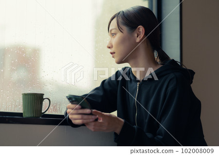 一位面容慵懶的年輕女子在窗邊喝咖啡,手裡拿著智能手機 一位面容慵懶的年輕女子在窗邊喝咖啡,手裡拿著智能手機 102008009