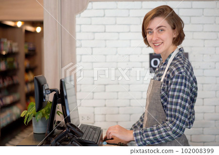 Caucasian white female shopkeeper standing behind a cashier counter and smiling to camera. Happy female shopkeeper or coffee shop staff looking and smiling at camera. 102008399