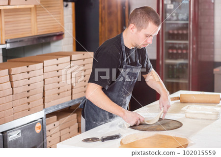 The chef prepares pizza. Raw pizza ready to bake. Cook in a blue apron in the kitchen. with a shovel in his hands. boxes for food delivery on background. The chef prepares pizza. Raw pizza ready to bake. Cook in a blue apron in the kitchen. with a shovel in his hands. boxes for food delivery on background. 102015559