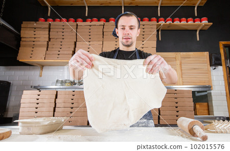 The chef prepares pizza. Raw pizza ready to bake. Cook in a blue apron in the kitchen. with a shovel in his hands. boxes for food delivery on background. The chef prepares pizza. Raw pizza ready to bake. Cook in a blue apron in the kitchen. with a shovel in his hands. boxes for food delivery on background. 102015576