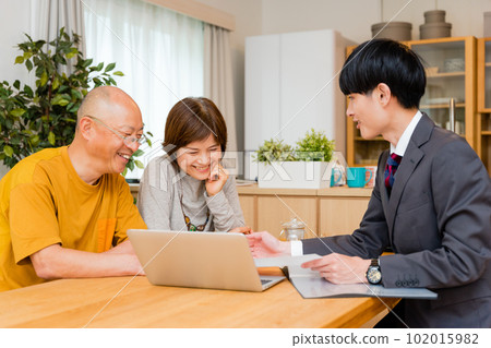 An elderly couple in their 50s and 60s listening to a young suit businessman explaining insurance products in the living room An elderly couple in their 50s and 60s listening to a young suit businessman explaining insurance products in the living room 102015982