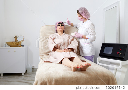 Cosmetologist applying black mask on the face of a beautiful woman for carbon peeling Cosmetologist applying black mask on the face of a beautiful woman for carbon peeling 102016142