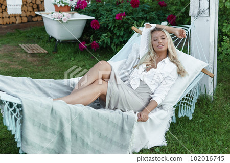 A beautiful blonde woman is lying on a hammock in her summer cottage. The concept of life in the country and summer holidays. A beautiful blonde woman is lying on a hammock in her summer cottage. The concept of life in the country and summer holidays. 102016745