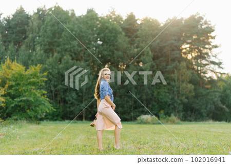 Young Caucasian woman or girl in a summer dress and a denim shirt is walking on the grass, holding shoes in her hands, relaxing outside the city on a weekend in summer 102016941