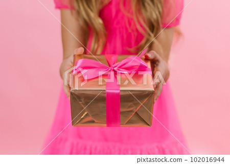 Girl is holding a golden gift with a pink fuchsia ribbon on a pink background. It's time to pack gifts. The concept of surprise is to surprise loved ones Girl is holding a golden gift with a pink fuchsia ribbon on a pink background. It's time to pack gifts. The concept of surprise is to surprise loved ones 102016944