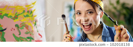 Joyful female artist smiling at camera, holding paintbrushes with her hands in paint while creating painting at home studio workshop Joyful female artist smiling at camera, holding paintbrushes with her hands in paint while creating painting at home studio workshop 102018138