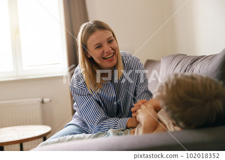 Overjoyed mother having fun with son at home, tickling and playing game on cozy couch in living room 102018352