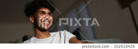 Cheerful young guy wearing pajamas taking some popcorn from a bowl while watching comedy, sitting on a couch at home Cheerful young guy wearing pajamas taking some popcorn from a bowl while watching comedy, sitting on a couch at home 102018428