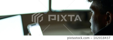 Close up shot of busy young guy sitting in dark room in front of many computer monitors and using smartphone while working alone late at night Close up shot of busy young guy sitting in dark room in front of many computer monitors and using smartphone while working alone late at night 102018437