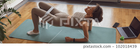 Slim woman doing exercises for legs with elastic band by online tutorials on laptop in living room Slim woman doing exercises for legs with elastic band by online tutorials on laptop in living room 102018439