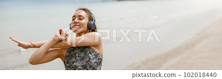 Beautiful young sportive woman in headphone making stretching exercises on beach. Blurred background Beautiful young sportive woman in headphone making stretching exercises on beach. Blurred background 102018440