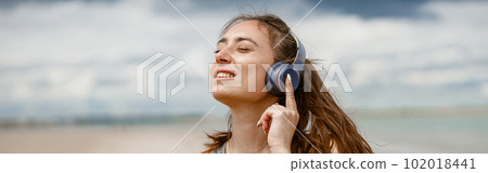 Smiling woman in sportswear listening music on headphones by the beach on background of ocean Smiling woman in sportswear listening music on headphones by the beach on background of ocean 102018441