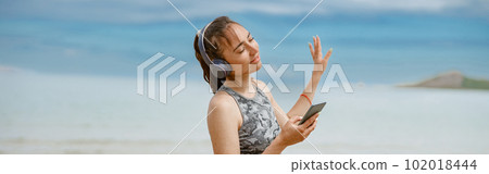 Woman in sportswear listening music on headphones by the beach on background of ocean Woman in sportswear listening music on headphones by the beach on background of ocean 102018444