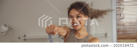 Slim woman doing exercises for hands with elastic band by online tutorials on laptop in living room 102018472