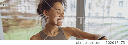 Smiling woman holding water bottle and looking at sports watch after exercise at home 102018473