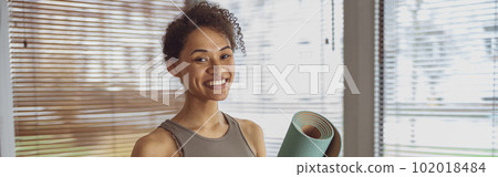 Woman holding rolled yoga or fitness mat and showing thumb up after working out at home 102018484