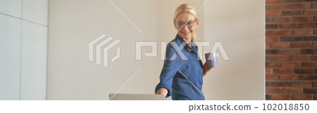 Cheerful mature businesswoman smiling, drinking coffee and using laptop computer while standing in modern kitchen room 102018850