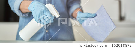 Close up shot of female hands in rubber gloves using detergent spray and cloth while cleaning kitchen surface 102018872