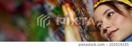 Close up portrait of relaxed young woman, female painter applying paint on canvas with fingers while creating a large modern abstract oil painting 102019285