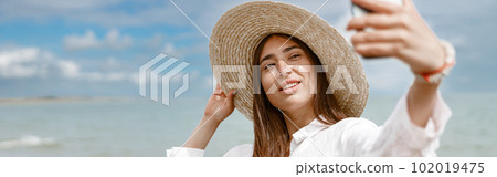 Beautiful woman in hat standing on beach near sea and making selphie Beautiful woman in hat standing on beach near sea and making selphie 102019475