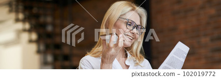 Business Approach. Stylish senior woman in glasses and business wear smiling, reading papers and talking on the phone, standing at home Business Approach. Stylish senior woman in glasses and business wear smiling, reading papers and talking on the phone, standing at home 102019772