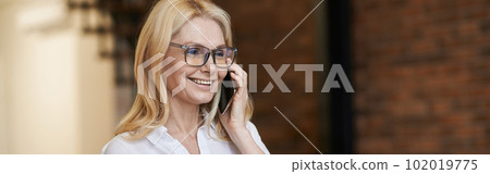 Morning. Cheerful middle aged woman with blonde hair and glasses holding a cup of coffee or tea while talking on her phone, standing at home Morning. Cheerful middle aged woman with blonde hair and glasses holding a cup of coffee or tea while talking on her phone, standing at home 102019775