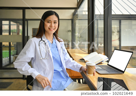 Confident asian female doctor in white coat and stethoscope using laptop in modern hospital canteen 102021649