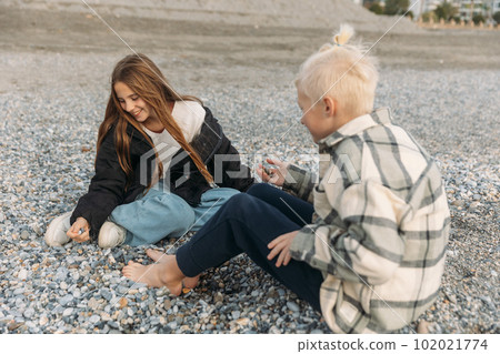 A young girl and a boy sit at seaside and chat, teen age friends talk, beautiful young girl laughs at her friend's jokes 102021774