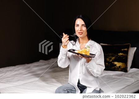Smiling brunette woman eating some cake in the bedroom in her apartment Smiling brunette woman eating some cake in the bedroom in her apartment 102022362