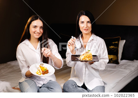Cheerful girls eating cakes, sitting on bedroom in apartment Cheerful girls eating cakes, sitting on bedroom in apartment 102022365