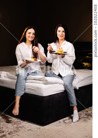 Cheerful girls eating cakes, sitting on bedroom in apartment Cheerful girls eating cakes, sitting on bedroom in apartment 102022408
