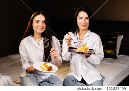 Cheerful girls eating cakes, sitting on bedroom in apartment Cheerful girls eating cakes, sitting on bedroom in apartment 102022409