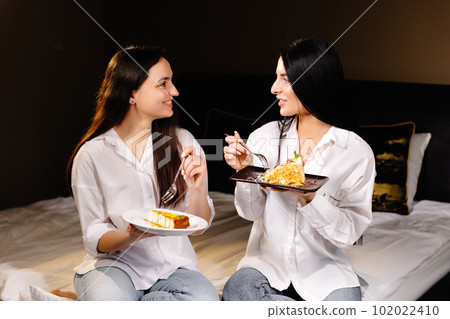 Cheerful girls eating cakes, sitting on bedroom in apartment Cheerful girls eating cakes, sitting on bedroom in apartment 102022410