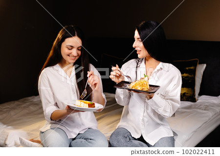 Cheerful girls eating cakes, sitting on bedroom in apartment 102022411