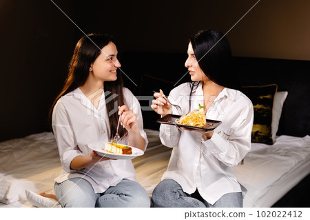 Cheerful girls eating cakes, sitting on bedroom in apartment Cheerful girls eating cakes, sitting on bedroom in apartment 102022412