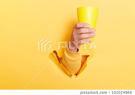Indoor shot of person hand breaking through yellow paper and holding cup with water or other liquid, showing mug with coffee or tea. 102024969