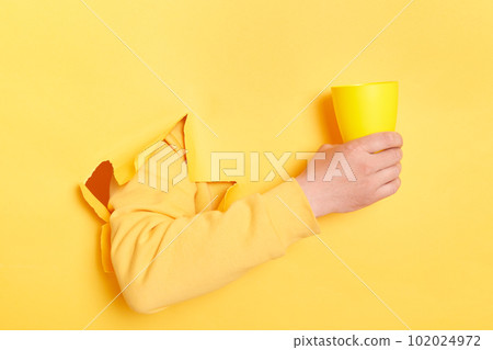 Human's holding in hand cup of coffee, tea or water isolated through torn yellow wall. Copy space advertisement place for text image promotional content Advertising area workspace mock up Human's holding in hand cup of coffee, tea or water isolated through torn yellow wall. Copy space advertisement place for text image promotional content Advertising area workspace mock up 102024972