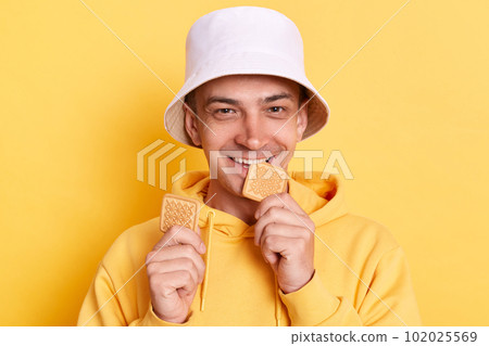 Hungry positive Caucasian young adult man wearing casual style hoodie and panama posing isolated over yellow background, holding and biting cookies, looking at camera with happiness. Hungry positive Caucasian young adult man wearing casual style hoodie and panama posing isolated over yellow background, holding and biting cookies, looking at camera with happiness. 102025569