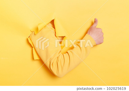 Indoor shot of human hand breaks arm through paper and showing a thumb up sign through a ripped hole, like gesture, isolated over yellow paper background. Indoor shot of human hand breaks arm through paper and showing a thumb up sign through a ripped hole, like gesture, isolated over yellow paper background. 102025619