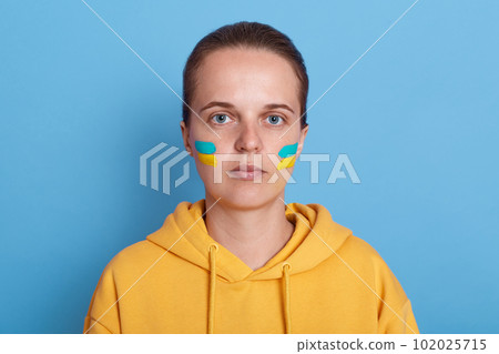 Portrait of sad serious woman wearing yellow hoodie and with flag of Ukraine on her cheek, looking at camera, expressing sorrow, isolated over blue background. War between Russia and Ukraine. 102025715