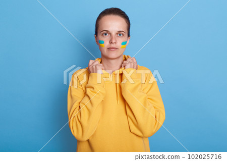 Indoor shot of serious Ukrainian woman wearing hoodie with blue and yellow flag on cheeks, holding hands on collar, looking at camera, support her motherland during war, isolated over blue background. 102025716
