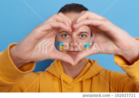 Portrait of woman wearing yellow hoodie showing love gesture with hands, making heart shape with her fingers, expressing love to Ukraine, supports during war against Russia. Glory to Ukraine. 102025721