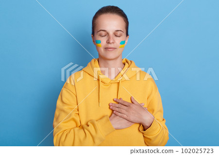 Portrait of woman wearing hoodie with blue and yellow flag on cheeks, keeps hands on heart, standing with closed eyes, thinking about Ukraine, war against Russia, posing isolated over blue background. Portrait of woman wearing hoodie with blue and yellow flag on cheeks, keeps hands on heart, standing with closed eyes, thinking about Ukraine, war against Russia, posing isolated over blue background. 102025723
