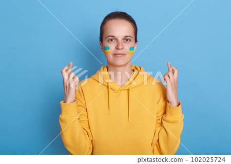 Adorable positive hopeful woman wearing hoodie with blue and yellow flag on cheeks, standing isolated over blue background and crossing fingers, making wish her country will free and win the war. Adorable positive hopeful woman wearing hoodie with blue and yellow flag on cheeks, standing isolated over blue background and crossing fingers, making wish her country will free and win the war. 102025724