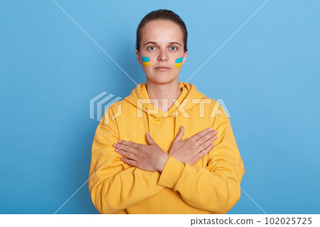 Portrait of serious woman wearing hoodie with blue and yellow flag on cheeks, isolated over blue background, keeps hands crossed on her chest, looking at camera. Glory to Ukraine. Portrait of serious woman wearing hoodie with blue and yellow flag on cheeks, isolated over blue background, keeps hands crossed on her chest, looking at camera. Glory to Ukraine. 102025725