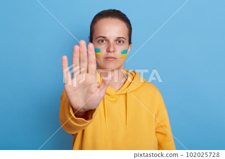 Indoor shot of serious woman in yellow hoodie with Ukrainian flag on cheeks, showing stop gesture with her palm, not war, peace in the world, posing isolated over blue background. 102025728