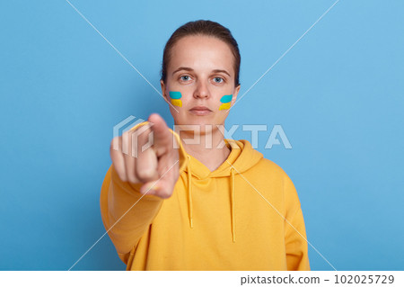 Indoor shot of serious patriotic woman wearing hoodie with blue and yellow flag on cheeks, looking and pointing finger to camera, Russian you can stop the war, posing isolated over blue background. 102025729