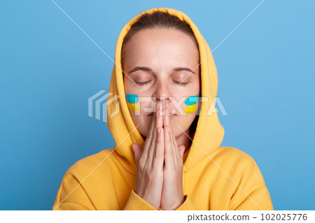 Portrait of sad hopeful woman wearing yellow hoodie and with flag of Ukraine on her cheek, praying for Ukraine people and stop of war, isolated over blue background. War between Russia and Ukraine. 102025776