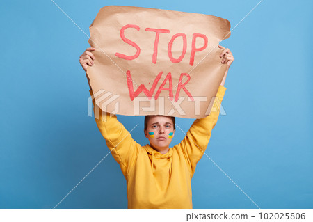 Sad woman holding Ukrainian flag and "Stop the War" placard sign in protest manifestation against Russia invasion on Ukraine. Russian attack, anti-war demonstration. Sad woman holding Ukrainian flag and "Stop the War" placard sign in protest manifestation against Russia invasion on Ukraine. Russian attack, anti-war demonstration. 102025806