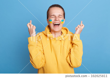 Portrait of happy woman wearing yellow hoodie and with flag of Ukraine on her cheek, standing with cross fingers, screaming, isolated over blue background. Glory to Ukraine. 102025811